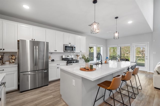 kitchen with appliances with stainless steel finishes, an island with sink, white cabinets, hanging light fixtures, and light hardwood / wood-style floors