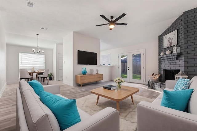 living room with a brick fireplace, ceiling fan with notable chandelier, light wood-type flooring, and french doors