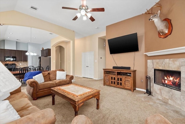 living room with ceiling fan with notable chandelier, light colored carpet, a tiled fireplace, and vaulted ceiling