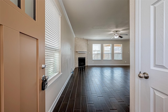 unfurnished living room with dark wood-style floors, ornamental molding, ceiling fan, a stone fireplace, and baseboards