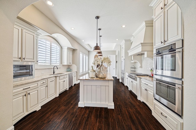 kitchen with premium range hood, a kitchen island, appliances with stainless steel finishes, hanging light fixtures, and light stone counters