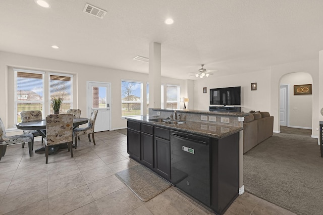 kitchen with sink, dishwasher, light colored carpet, dark stone counters, and a kitchen island with sink