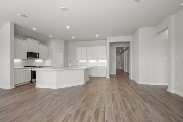 kitchen featuring appliances with stainless steel finishes, tasteful backsplash, white cabinets, a center island with sink, and light wood-type flooring