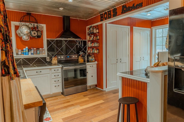 kitchen featuring stainless steel electric stove, wood counters, white cabinets, light hardwood / wood-style floors, and wall chimney range hood