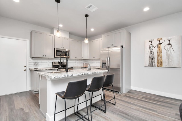 kitchen featuring hanging light fixtures, stainless steel appliances, light stone counters, tasteful backsplash, and an island with sink