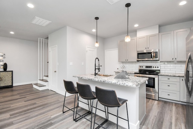 kitchen featuring decorative light fixtures, an island with sink, sink, light stone counters, and stainless steel appliances