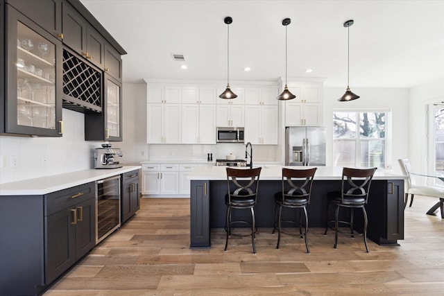 kitchen with hanging light fixtures, appliances with stainless steel finishes, an island with sink, beverage cooler, and white cabinets