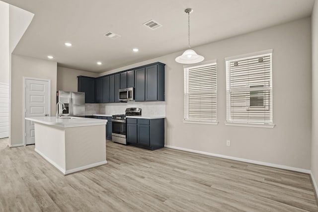 kitchen with pendant lighting, appliances with stainless steel finishes, an island with sink, and light hardwood / wood-style flooring