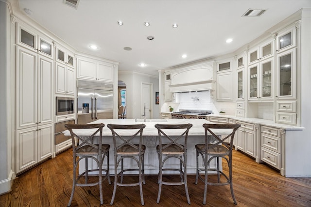 kitchen with white cabinetry, an island with sink, premium range hood, and built in appliances