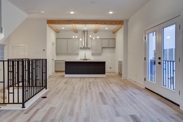 kitchen with wall chimney range hood, gray cabinets, a kitchen island with sink, light hardwood / wood-style floors, and french doors