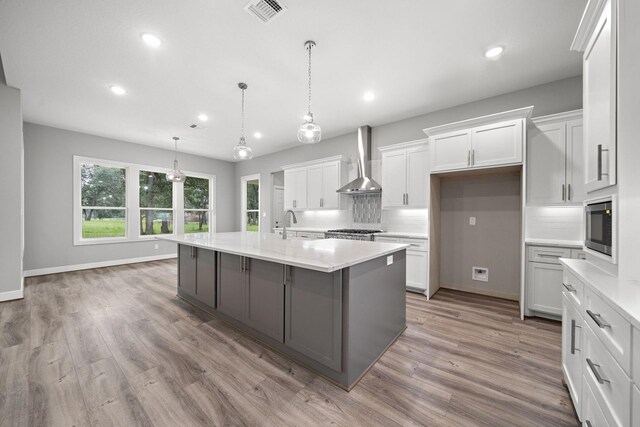 kitchen with visible vents, hanging light fixtures, a kitchen island with sink, wall chimney range hood, and white cabinetry