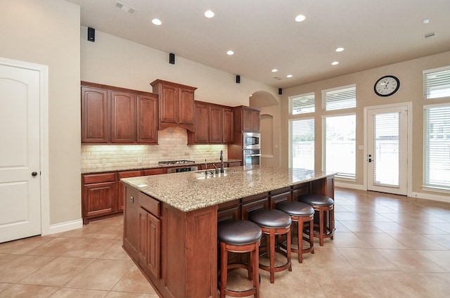 kitchen featuring sink, backsplash, light stone counters, an island with sink, and a healthy amount of sunlight