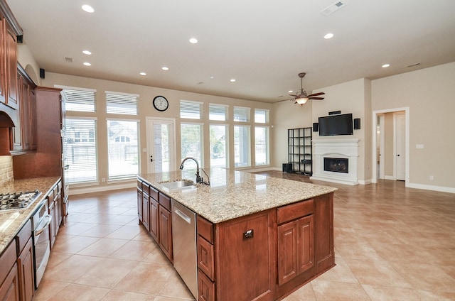 kitchen featuring sink, ceiling fan, stainless steel appliances, light stone countertops, and a center island with sink