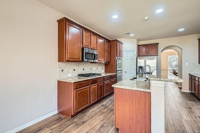 kitchen with sink, an island with sink, stainless steel appliances, light stone countertops, and decorative backsplash