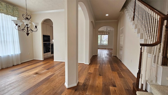 hallway with arched walkways, wood finished floors, stairs, ornamental molding, and an inviting chandelier