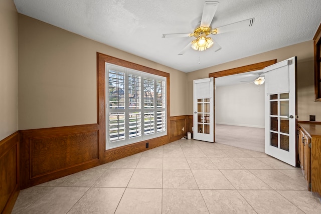 empty room featuring french doors, light tile patterned floors, a ceiling fan, wainscoting, and a textured ceiling