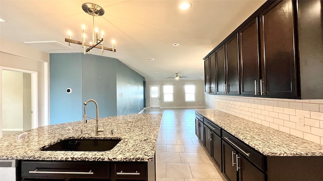 kitchen with sink, light stone countertops, dark brown cabinetry, a kitchen island with sink, and tasteful backsplash