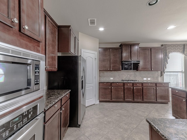 kitchen featuring dark brown cabinets, dark stone countertops, light tile patterned floors, stainless steel appliances, and backsplash