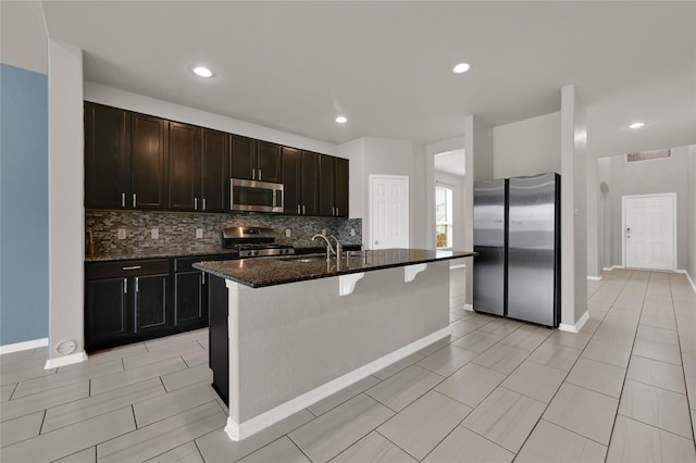 kitchen with sink, appliances with stainless steel finishes, an island with sink, dark stone counters, and backsplash