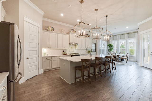 kitchen with hanging light fixtures, backsplash, stainless steel appliances, an island with sink, and white cabinets