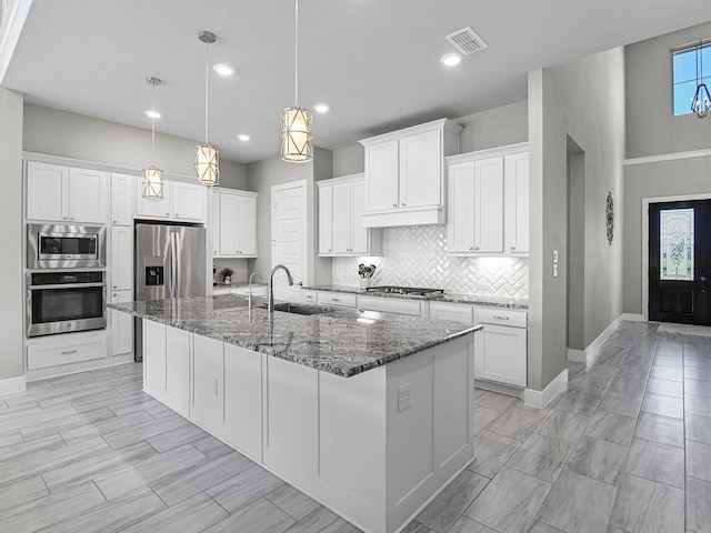 kitchen with appliances with stainless steel finishes, sink, dark stone countertops, white cabinets, and a kitchen island with sink