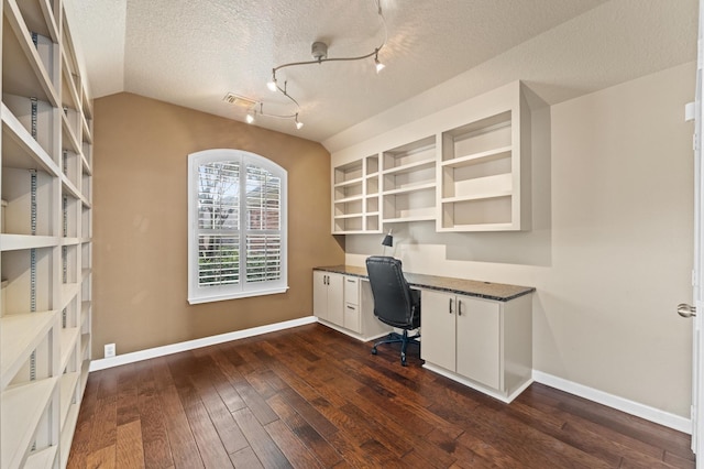 unfurnished office featuring rail lighting, built in desk, vaulted ceiling, a textured ceiling, and dark hardwood / wood-style floors