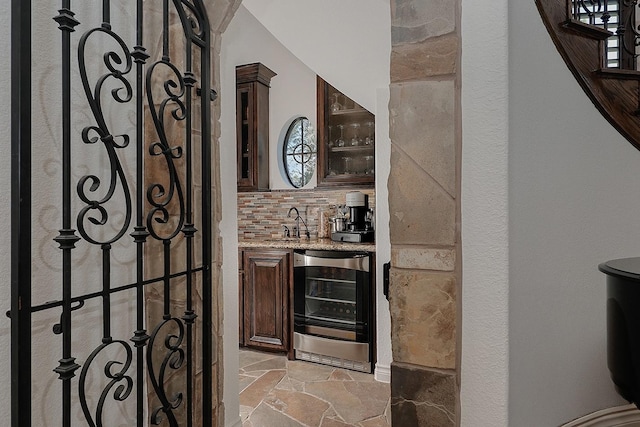 bar featuring wine cooler, dark brown cabinetry, sink, light stone counters, and decorative backsplash