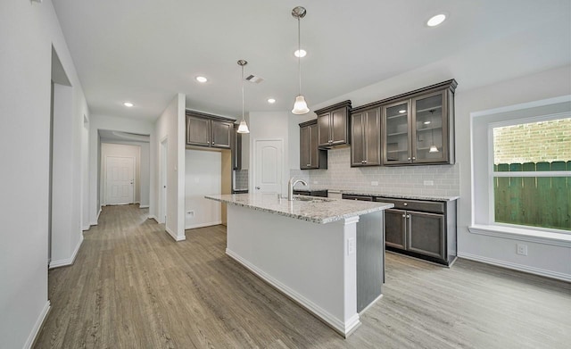 kitchen with sink, hanging light fixtures, dark brown cabinetry, light stone countertops, and an island with sink