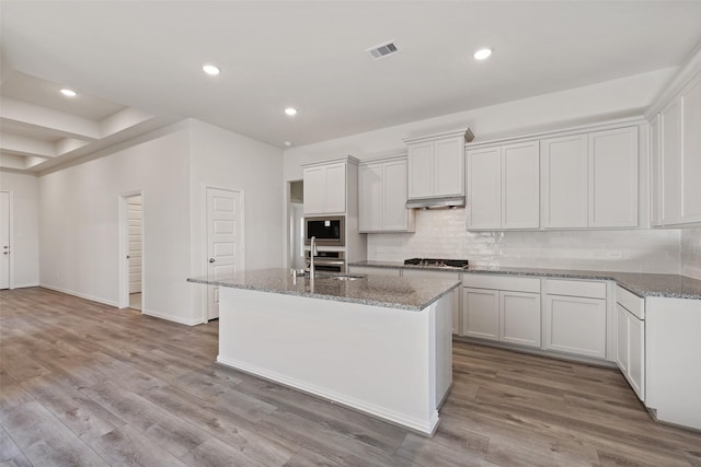 27603 Beachside Arbor Dr, Katy, TX 77493 - kitchen featuring stainless steel appliances, a kitchen island with sink, under cabinet range hood, backsplash, and wood finished floors