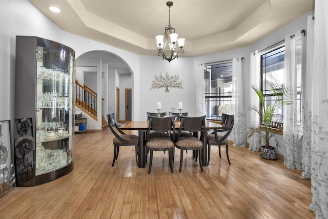 dining space with arched walkways, an inviting chandelier, stairs, a tray ceiling, and light wood-type flooring