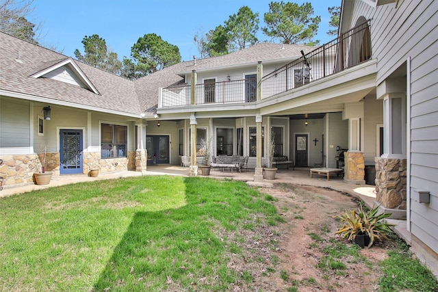 exterior space with a balcony, a shingled roof, stone siding, a lawn, and a patio area