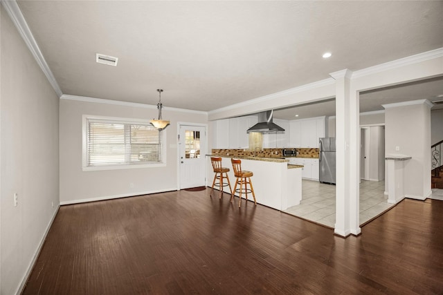 kitchen with wall chimney range hood, appliances with stainless steel finishes, white cabinetry, hanging light fixtures, and a kitchen bar