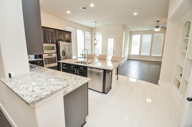 kitchen with sink, light stone counters, hanging light fixtures, appliances with stainless steel finishes, and an island with sink