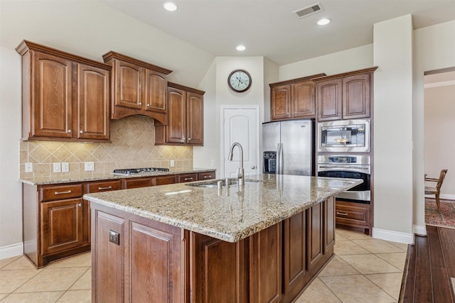 kitchen with light tile patterned flooring, sink, light stone counters, stainless steel appliances, and a kitchen island with sink