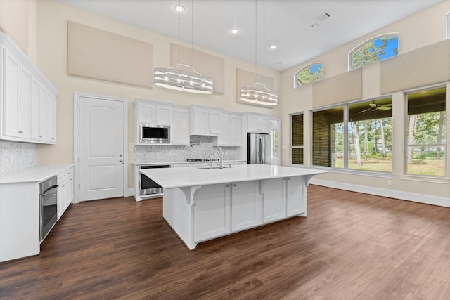 kitchen featuring hanging light fixtures, a towering ceiling, stainless steel appliances, a kitchen island with sink, and white cabinets