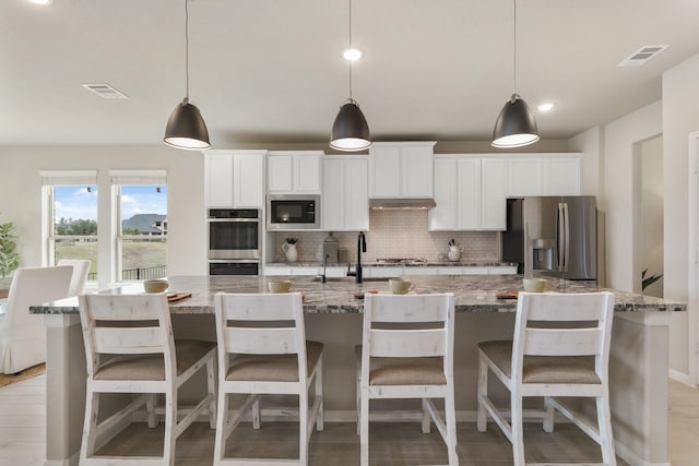 kitchen with tasteful backsplash, decorative light fixtures, a large island with sink, appliances with stainless steel finishes, and light stone countertops