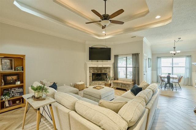 living room with light parquet floors, a tray ceiling, a fireplace, ornamental molding, and ceiling fan with notable chandelier