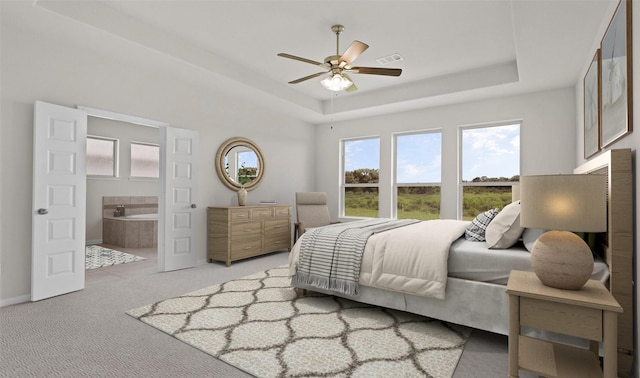 bedroom with a raised ceiling, ensuite bathroom, light colored carpet, and ceiling fan