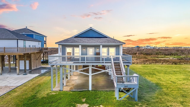 back of property at dusk with covered porch, a shingled roof, stairs, driveway, and a lawn