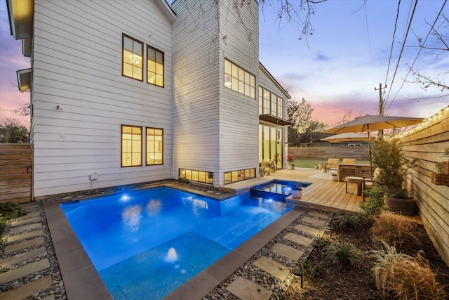 pool at dusk featuring a fenced backyard, a wooden deck, and a pool with connected hot tub