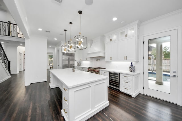 kitchen with wine cooler, custom exhaust hood, light countertops, glass insert cabinets, and a kitchen island with sink