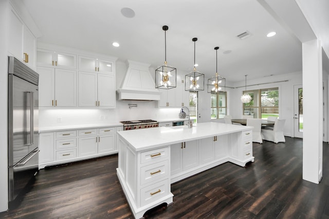 kitchen featuring glass insert cabinets, decorative light fixtures, a kitchen island with sink, custom exhaust hood, and built in fridge