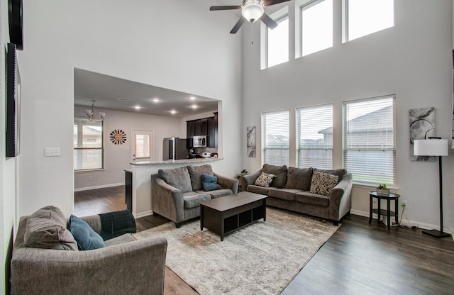 living room featuring dark wood-type flooring, a towering ceiling, a healthy amount of sunlight, and ceiling fan with notable chandelier