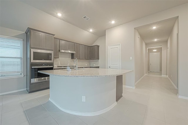 kitchen with appliances with stainless steel finishes, sink, decorative backsplash, light stone countertops, and a center island with sink