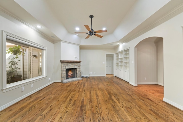 unfurnished living room featuring a stone fireplace, hardwood / wood-style flooring, ceiling fan, a raised ceiling, and built in shelves