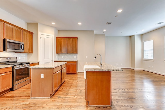 kitchen featuring decorative backsplash, a kitchen island with sink, a sink, light wood-style flooring, and appliances with stainless steel finishes