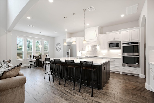 kitchen with built in appliances, decorative light fixtures, custom exhaust hood, and a center island with sink