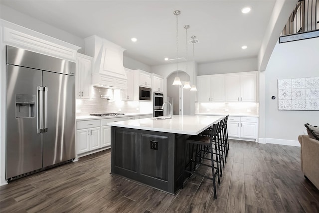 kitchen with premium range hood, white cabinetry, an island with sink, hanging light fixtures, and built in appliances