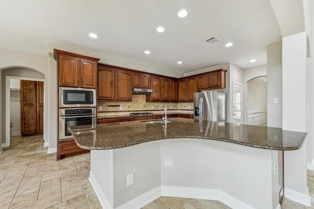 kitchen with sink, appliances with stainless steel finishes, dark stone countertops, tasteful backsplash, and a center island with sink