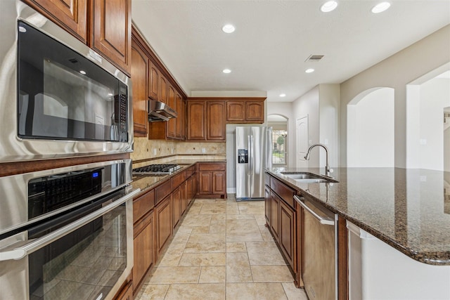 kitchen with sink, dark stone countertops, appliances with stainless steel finishes, an island with sink, and backsplash